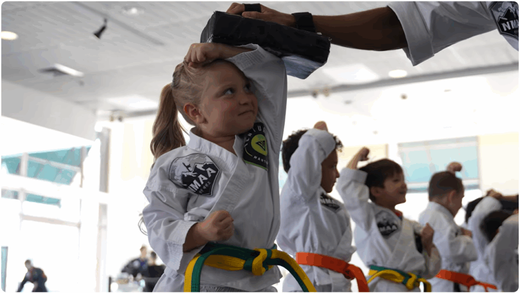 Young girl in a martial arts uniform practicing a technique with a training pad held above her head.