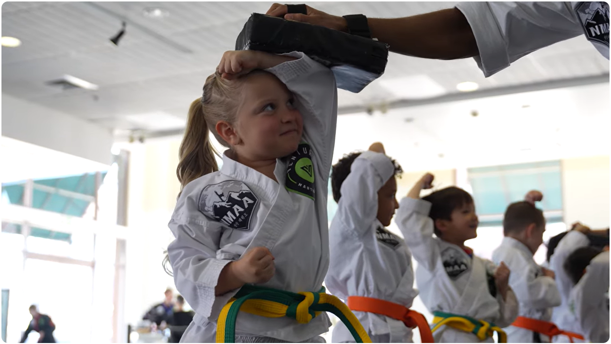 Young girl in a martial arts uniform practicing a technique with a training pad held above her head.