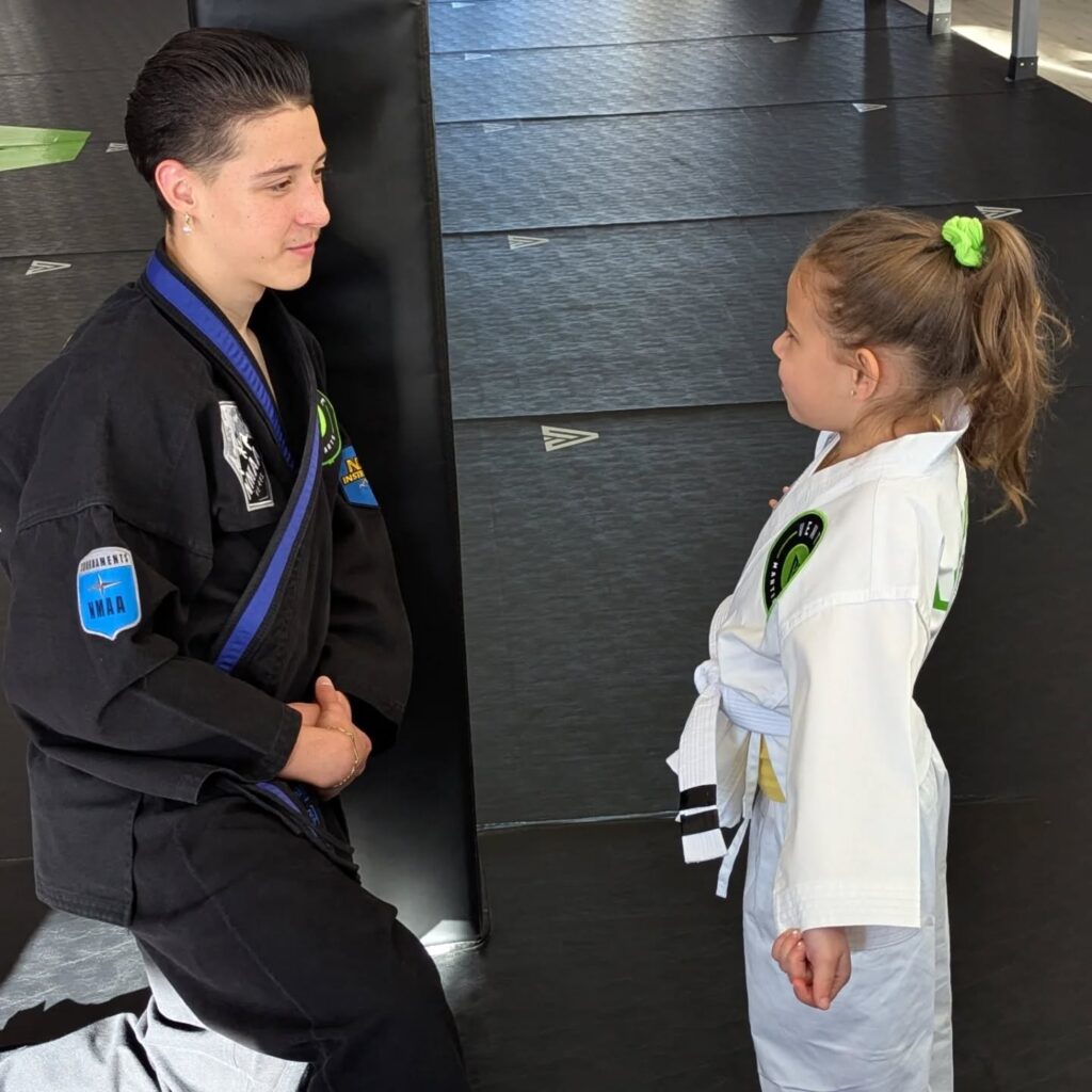 A young martial arts instructor kneels and engages in conversation with a child in a white karate uniform during a training session on a black mat.