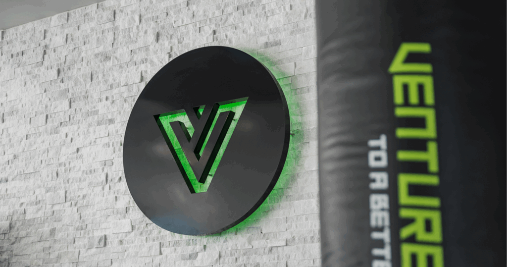 Logo of Venture Martial Arts displayed on a textured wall, featuring a black circular design with a green accent.