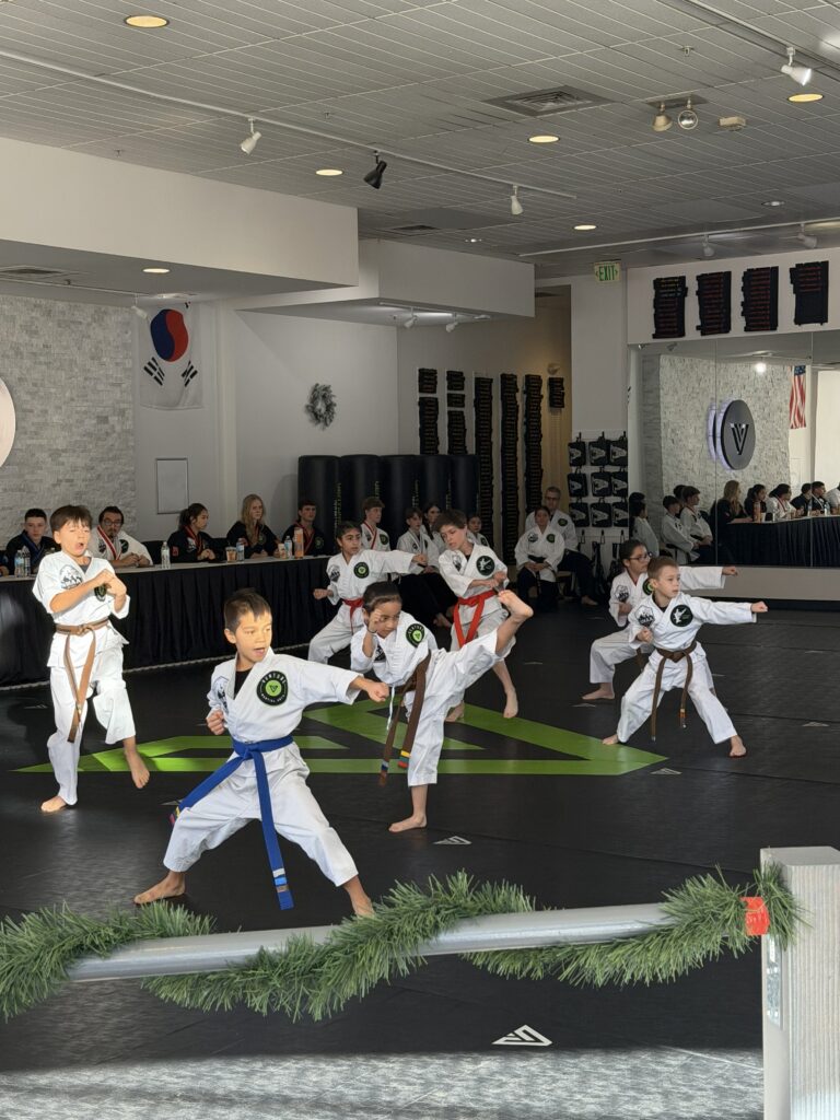 Children practicing martial arts in a dojo, showcasing various techniques and forms during a training session, with an audience observing in the background.