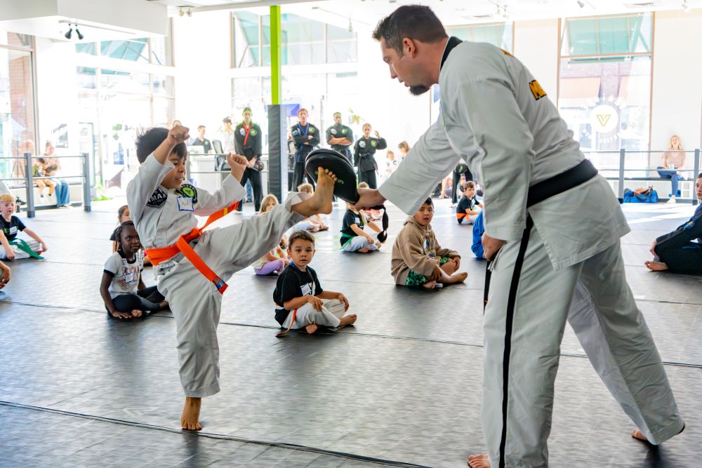 A child wearing an orange belt performs a kick while training in a martial arts studio, with an instructor holding a pad.
