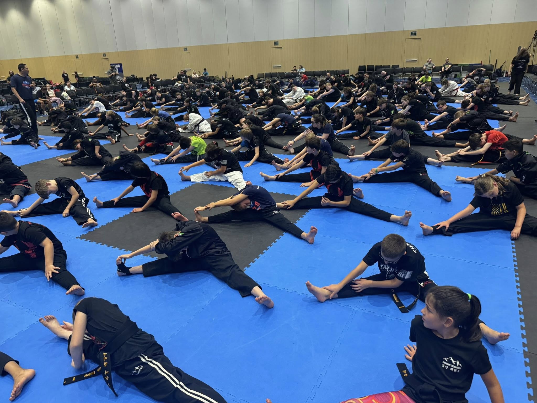 Large group of martial arts students practicing stretches on blue mats in a spacious training facility.