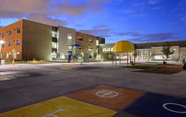 Swigert International School building at dusk featuring a playground, colorful play area markings, and well-lit classrooms, surrounded by landscaped greenery.